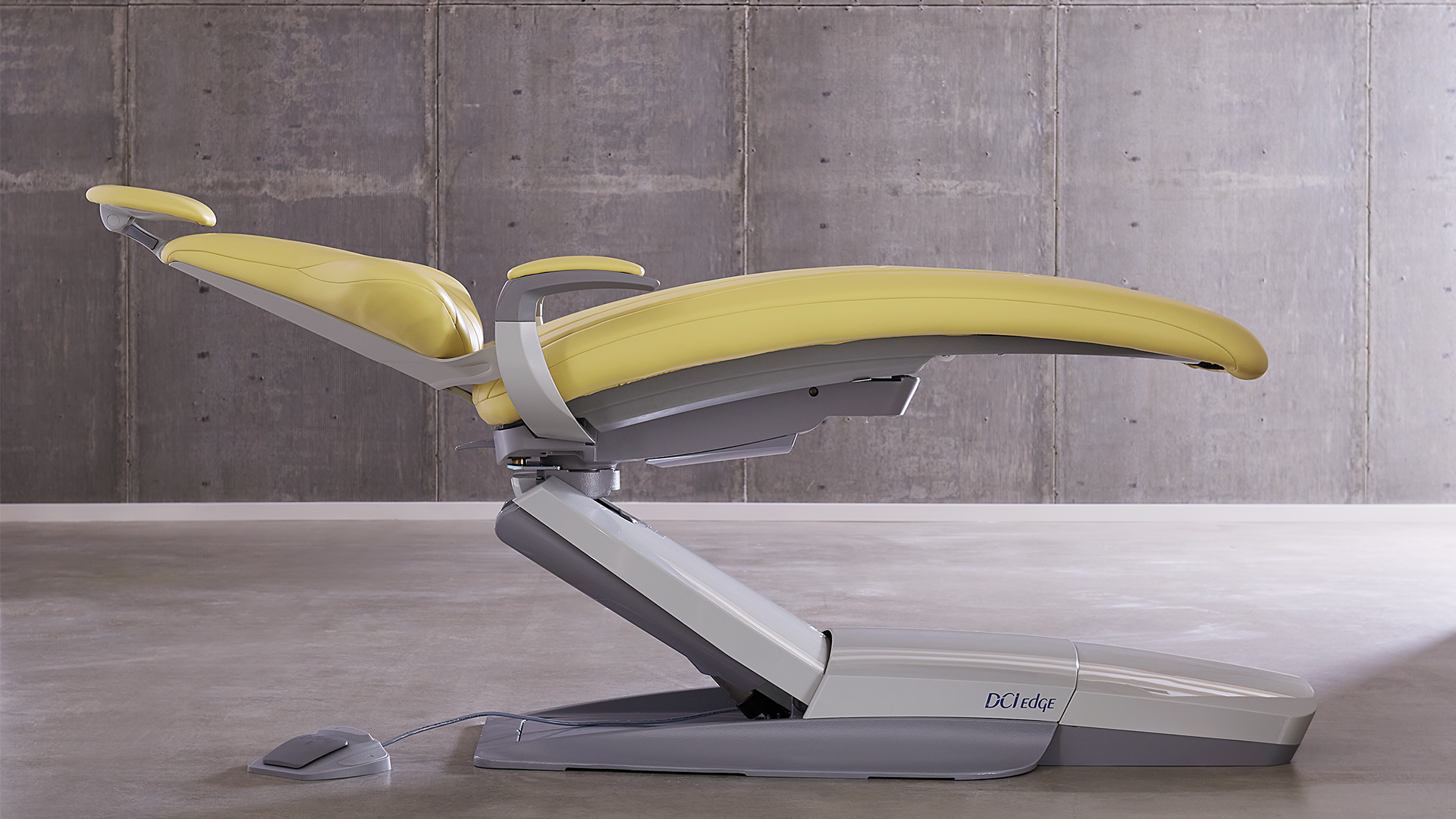 Yellow reclining dental chair with grey base positioned against a concrete wall background.